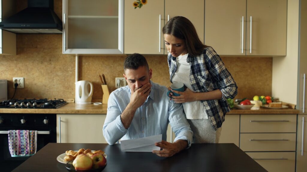 Upset young man reading unpaid bills and hugged by his wife supporting him early morning in the kitchen at home