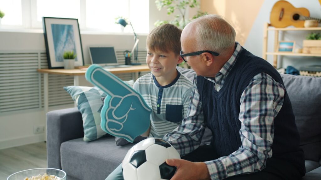 Joyful family grandfather and grandson are watching football on TV cheering hugging celebrating victory. Sports fans, modern lifestyle and leisure concept.