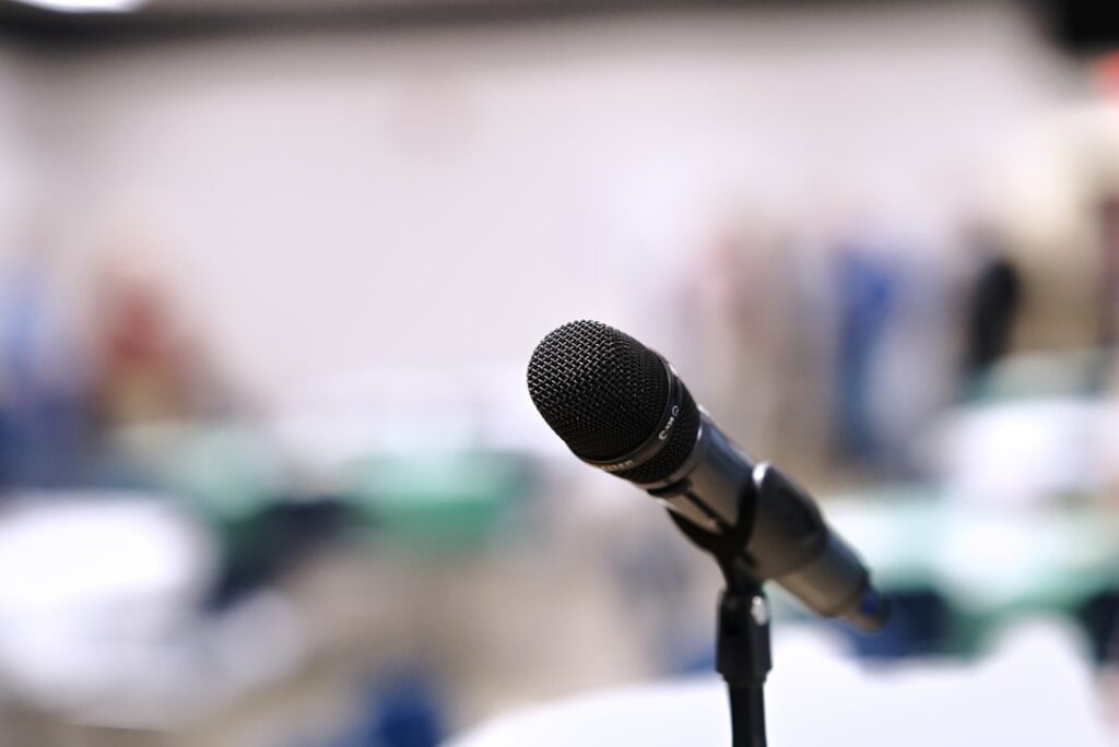 Microphone in focus in front of a room full of tables and people standing awaiting a speech