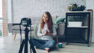Happy female blogger is recording video for internet videolog talking and gesturing showing thumbs-up and waving hand. Blogging, young people and success concept.