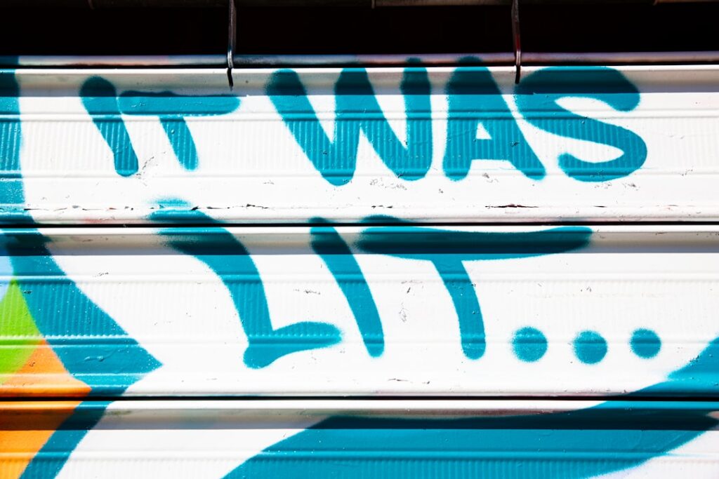 A bright blue graffiti message sprayed across a white metal storefront shutter reads: “IT WAS LIT....” The oversized lettering and trailing dots capture the fleeting language of nightlife and memory, freezing a casual street expression into an urban time capsule.
