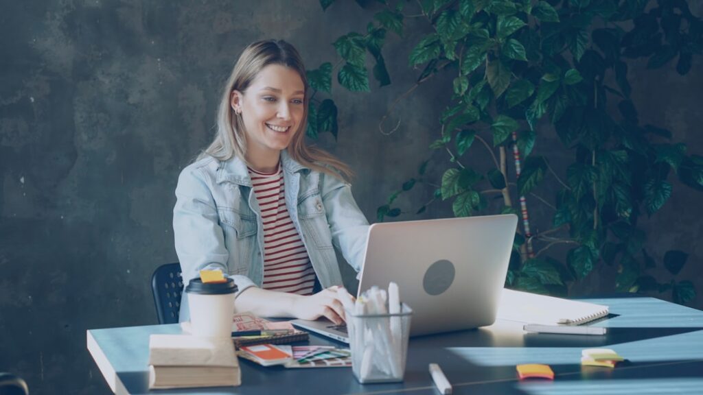 Attractive young woman in casual clothes is talking on skype on laptop while sitting at table in office. She is speaking emotionally, gesturing and laughing. Modern communication concept.