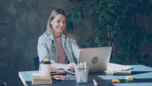 Attractive young woman in casual clothes is talking on skype on laptop while sitting at table in office. She is speaking emotionally, gesturing and laughing. Modern communication concept.