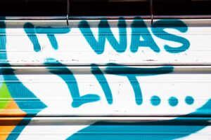 A bright blue graffiti message sprayed across a white metal storefront shutter reads: “IT WAS LIT....” The oversized lettering and trailing dots capture the fleeting language of nightlife and memory, freezing a casual street expression into an urban time capsule.