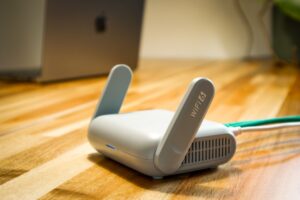 A stylish, compact WiFi 6 router with dual antennas stands prominently on a wooden desk, illuminated by a subtle blue LED. Connected via green Ethernet and white power cables, this modern device promises fast and reliable wireless connectivity. The softly blurred background hints at a contemporary workspace, with a laptop partially visible, making this image perfect for illustrating advanced home networking, tech upgrades, or smart office environments.