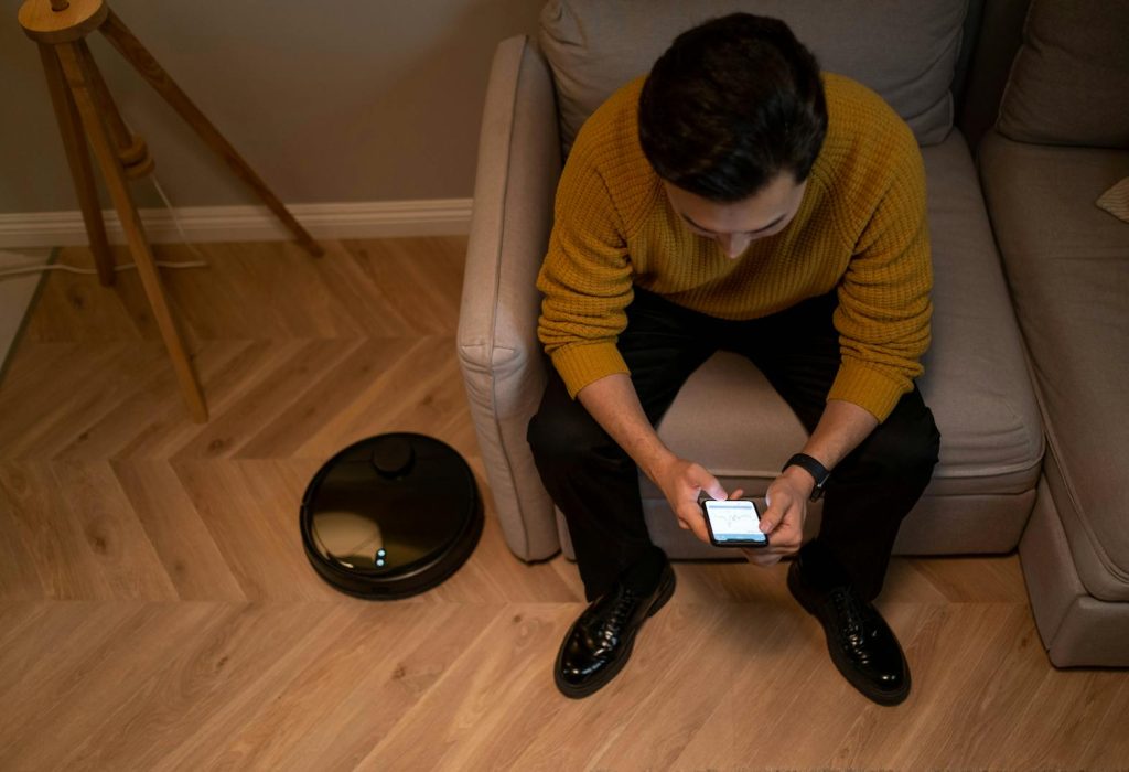 Man sits on sofa using a smartphone, next to a robotic vacuum cleaner in a modern living room.